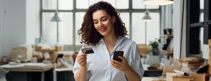 Mulher com um celular e um cartão nas mãos, finalizando uma compra online.